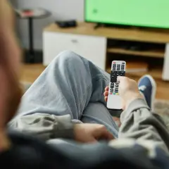 Osoba z pilotem w ręku, zastanawia się, kto nie musi płacić abonamentu RTV, oglądając zielony ekran telewizora.