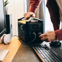 Mężczyzna podłącza wtyczkę do przenośnej stacji zasilania ATX, obok monitora i słuchawek.