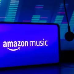 Słuchawki wiszą obok smartfona z logo Amazon Music. Tło to niebieski equalizer, sugerujący muzyczną przygodę z Amazon Music Polska.