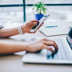 Kobieta trzyma telefon, planując, jak udostępnić internet z telefonu na komputer. Pracuje przy laptopie.