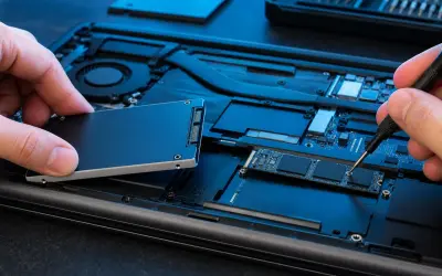 Wymiana dysku w laptopie: ręka trzyma nowy dysk SSD, druga dokręca śrubkę precyzyjnym śrubokrętem.