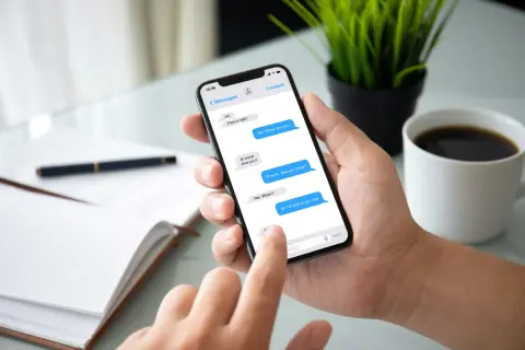 Dłoń trzyma smartfon z otwartym komunikatorem. Na ekranie widać rozmowę: "At work. See you today?". Obok filiżanka kawy.