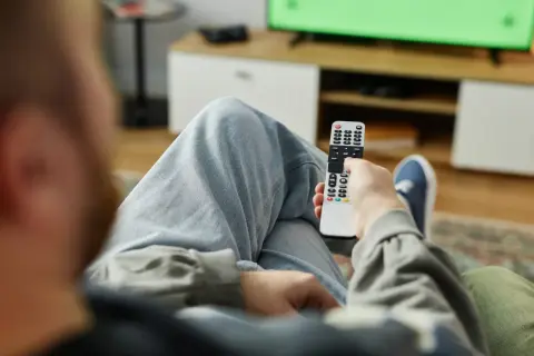 Osoba z pilotem w ręku, zastanawia się, kto nie musi płacić abonamentu RTV, oglądając zielony ekran telewizora.