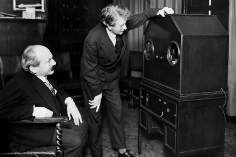 Dwóch mężczyzn przy pierwszym telewizorze. Jeden siedzi, drugi stoi i dotyka obudowy.