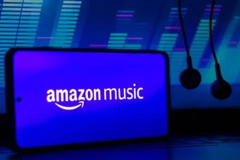 Słuchawki wiszą obok smartfona z logo Amazon Music. Tło to niebieski equalizer, sugerujący muzyczną przygodę z Amazon Music Polska.