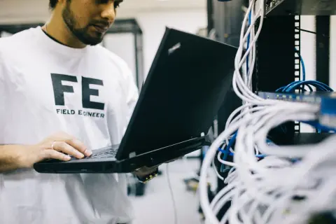 Inżynier w białej koszulce z napisem "FE FIELD ENGINEER" sprawdza połączenia sieciowe na laptopie, analizując ruch icmp.