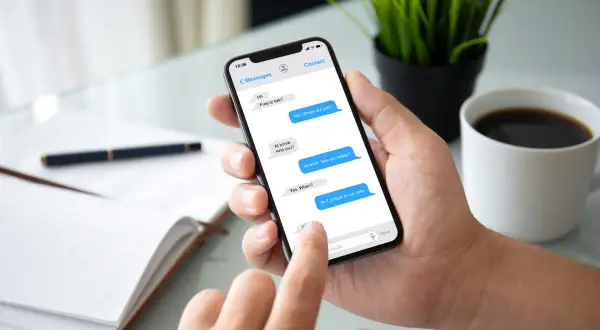 Dłoń trzyma smartfon z otwartym komunikatorem. Na ekranie widać rozmowę: "At work. See you today?". Obok filiżanka kawy.