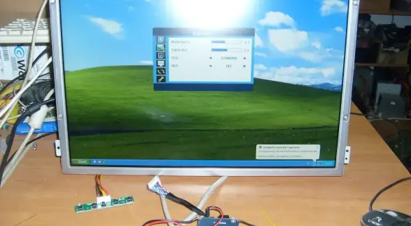 Na biurku leży monitor z pulpitem Windows XP, obok kable i płytka sterująca. To może być pomocne, jak podłączyć monitor do laptopa.