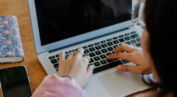 Kobieta pisze na laptopie. Obok leży telefon i notatnik. To jest print screen z jej pracy.