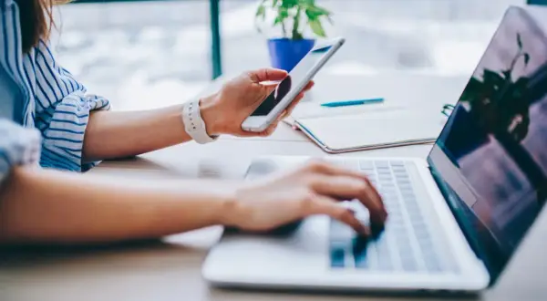 Kobieta trzyma telefon, planując, jak udostępnić internet z telefonu na komputer. Pracuje przy laptopie.
