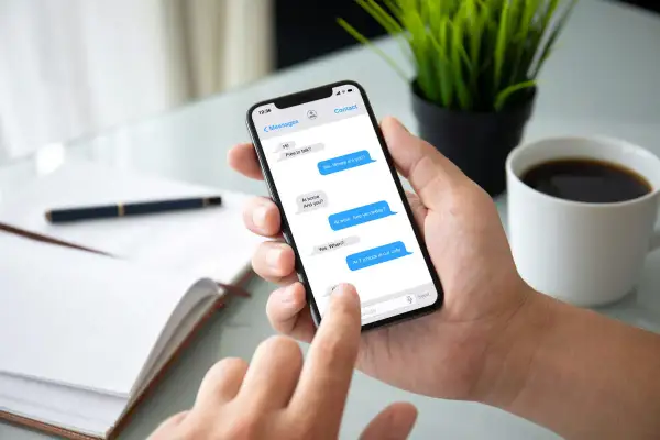 Dłoń trzyma smartfon z otwartym komunikatorem. Na ekranie widać rozmowę: "At work. See you today?". Obok filiżanka kawy.