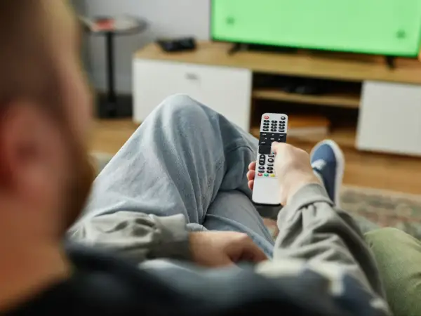 Osoba z pilotem w ręku, zastanawia się, kto nie musi płacić abonamentu RTV, oglądając zielony ekran telewizora.